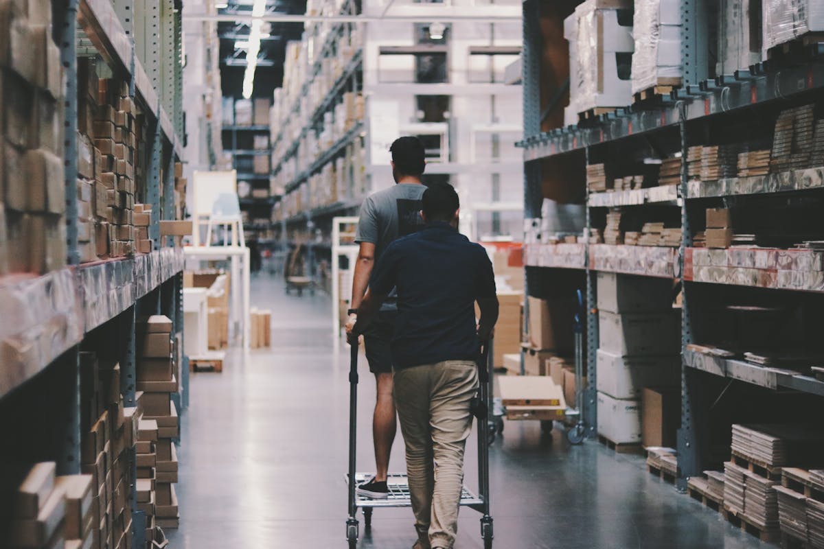 Como Planejar o Estoque para a Temporada de Vendas de Final de Ano Two men maneuver a trolley in a large warehouse filled with boxes and shelves.