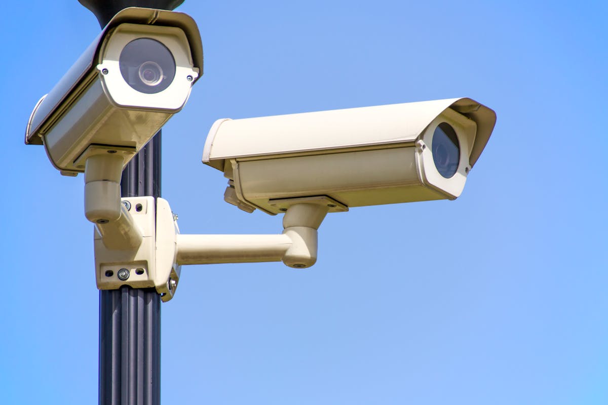 Qual a importância da segurança patrimonial nos dias atuais? Outdoor security cameras mounted on a pole against a clear blue sky, ensuring vigilant surveillance.