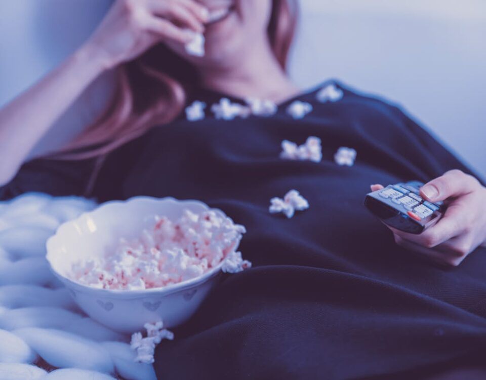 Por que assistir filmes pode melhorar seu humor? Woman relaxing on couch eating popcorn while watching TV, holding remote control, indoors.