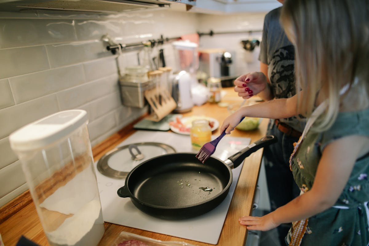 Qual é o segredo das receitas que derretem na boca? Young girl cooking with a frying pan under adult supervision in a modern kitchen.