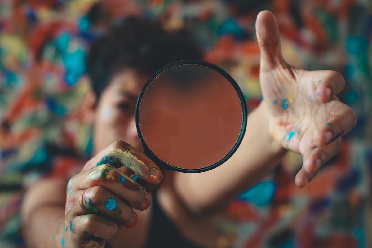 Como a curiosidade molda nossos desejos pessoais Artist with painted hands holding a lens in front of vibrant abstract background.