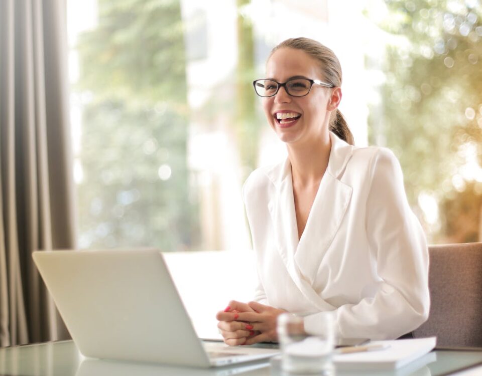 Automação comercial: O que realmente pode otimizar? Cheerful businesswoman in glasses working on a laptop, in a bright and modern office setting.