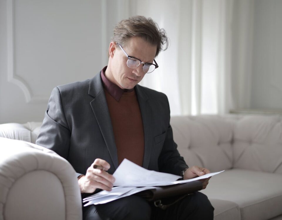 Como escolher entre seguro e cobertura médica ideal? Elegant adult man in jacket and glasses looking through documents while sitting on white sofa in luxury room
