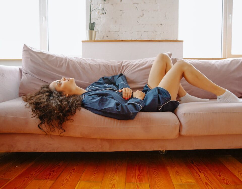 Quais os mitos mais comuns sobre doenças inflamatórias? A woman in casual wear experiences abdominal discomfort while lying on a pink couch indoors.