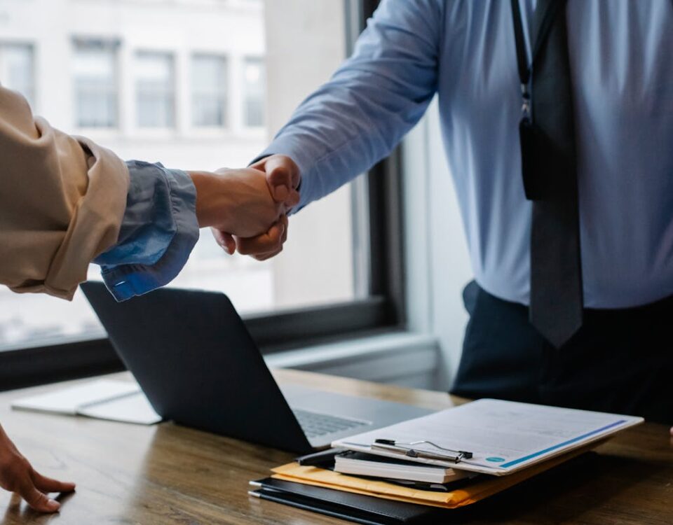 Vale a pena investir em seguro de vida hoje? Crop unrecognizable coworkers in formal wear standing at table with laptop and documents while greeting each other before meeting