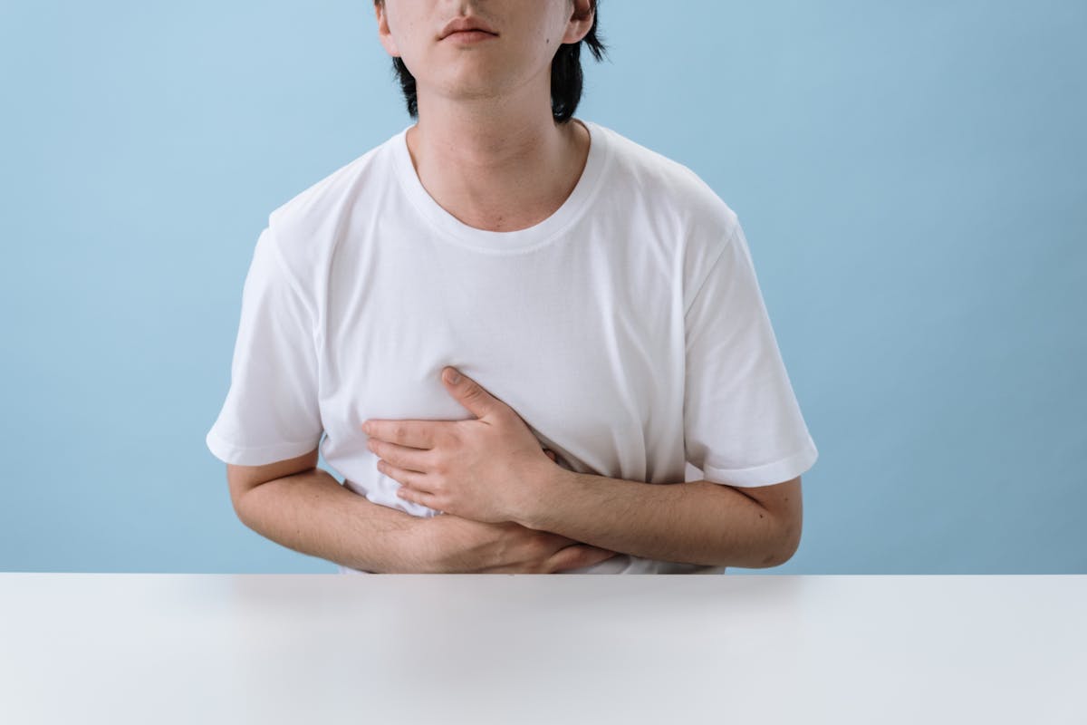 Quais os mitos mais comuns sobre doenças inflamatórias? An adult man in a white shirt holds his stomach, indicating pain and discomfort, with a light blue background.