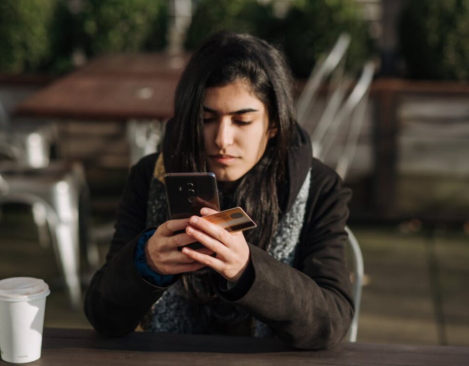 O que significa CVV? A pergunta mais comum sobre cartões Young ethnic female purchaser with credit card shopping online on mobile phone at table with takeaway hot drink in cafeteria