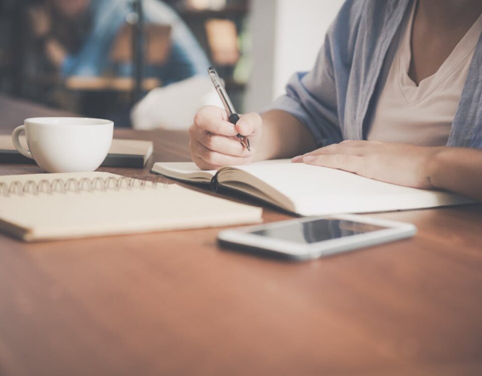 Quais profissões têm o futuro mais promissor no Brasil? A woman writes in a notebook at a café table with a coffee and smartphone nearby.