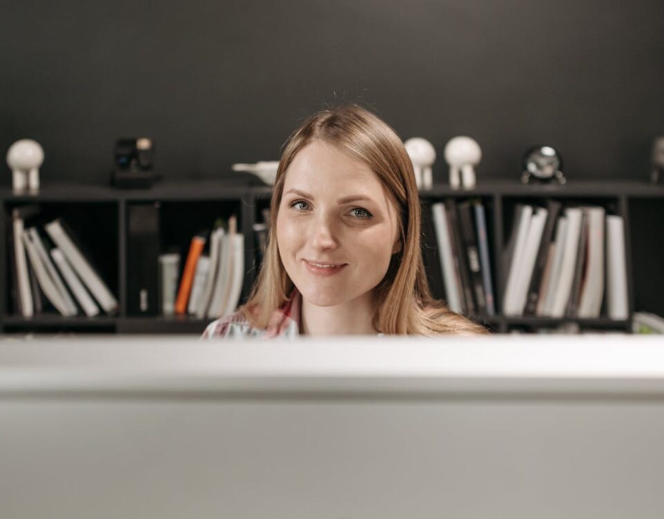 Qual é o futuro do auxiliar administrativo? Friendly young woman at her desk with bookshelves in modern office setting.