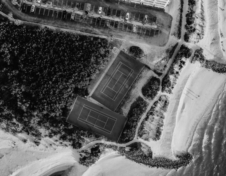 Como otimizar o uso de arenas esportivas? Aerial view of tennis courts near a beach, captured in black and white.