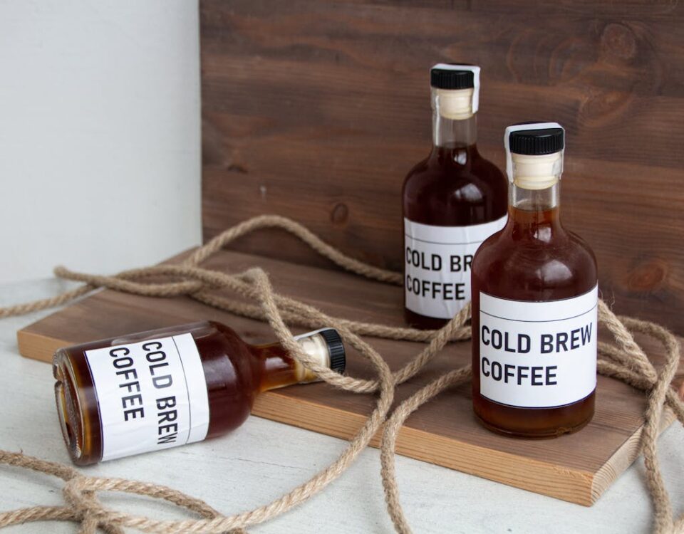 Qual é o segredo por trás dos rótulos adesivos perfeitos? Three bottles of cold brew coffee arranged artistically on wooden boards with rope accents, conveying a rustic vibe.