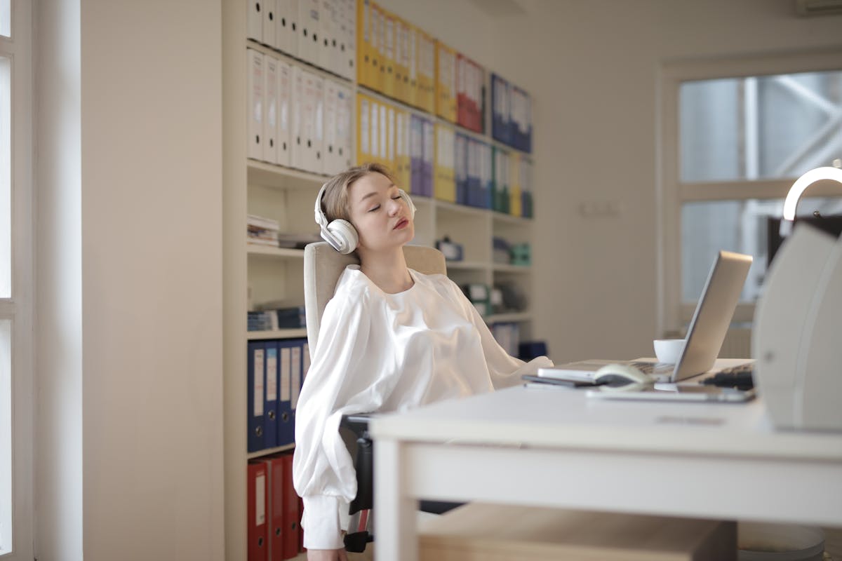 Por que certos sons parecem mais agradáveis? Woman in white shirt relaxing at office desk with headphones, appearing restful.