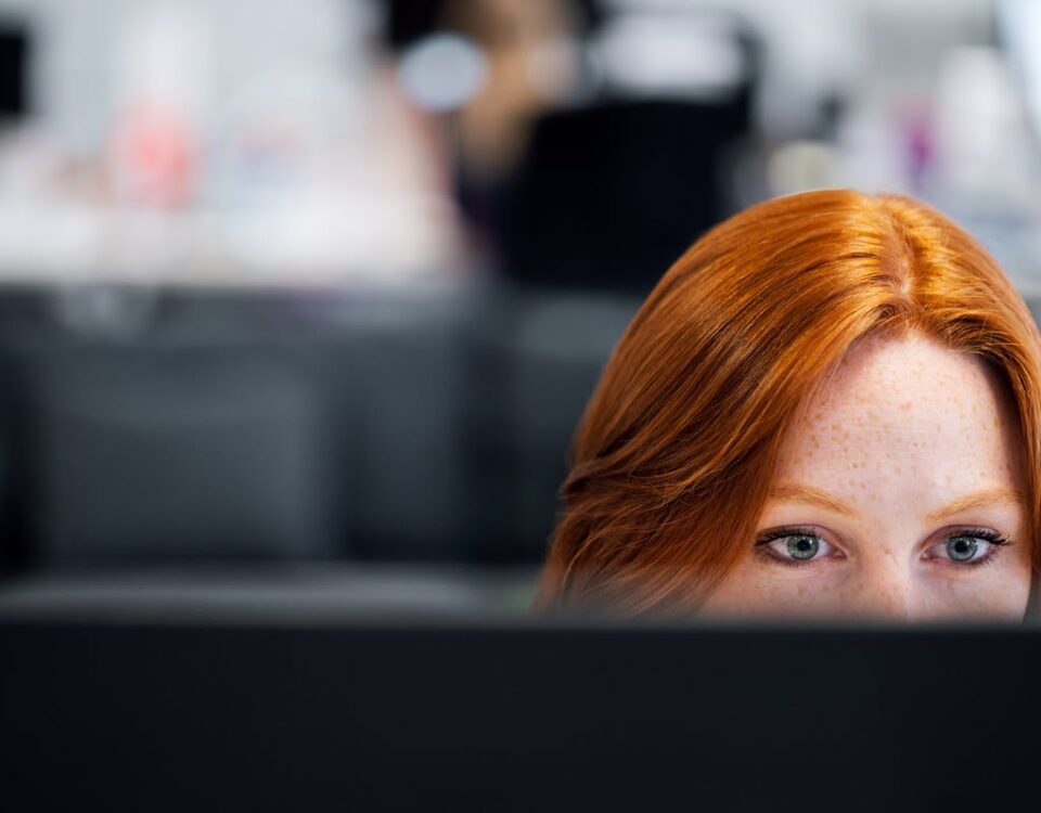Qual o segredo das empresas de TI mais inovadoras? A woman with red hair intensely focused on a computer screen in an office setting.