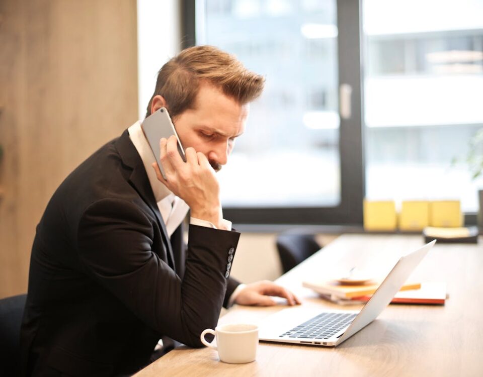 Como otimizar o atendimento sem aumentar os custos? A businessman in a suit making a phone call while working on a laptop in a modern office setting.