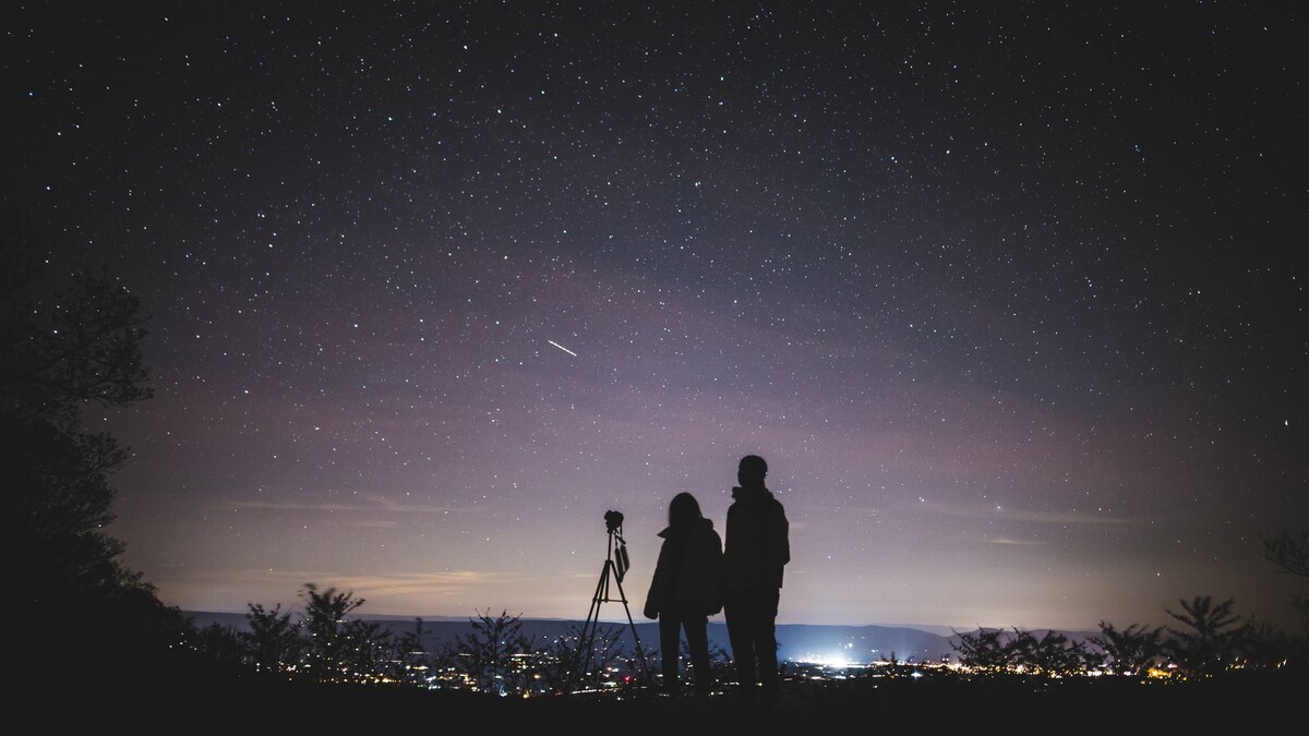 Silhuetas de duas pessoas com tripé observando um céu estrelado sobre uma cidade iluminada ao longe