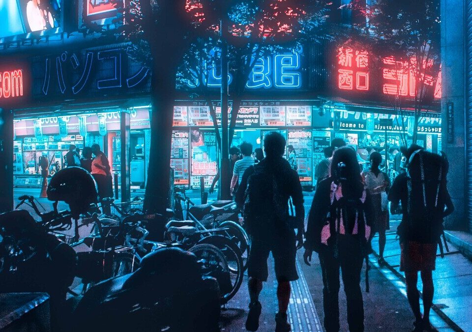 Rua urbana iluminada à noite, com pessoas caminhando e fachadas comerciais.