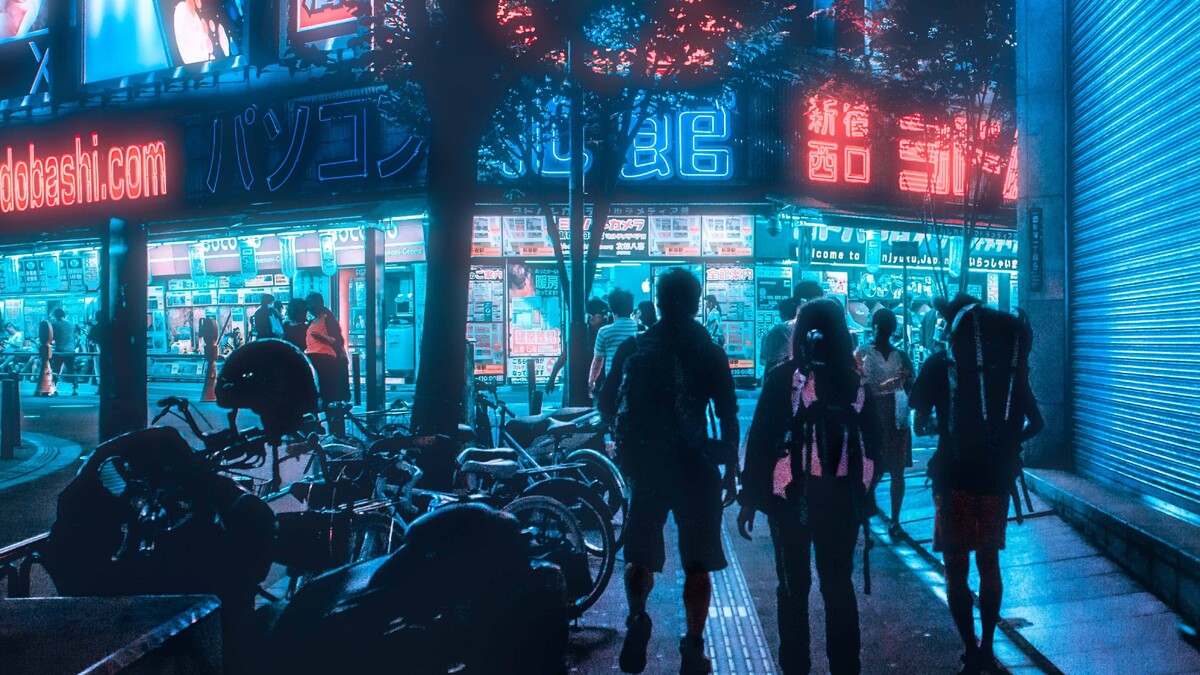 Rua urbana iluminada à noite, com pessoas caminhando e fachadas comerciais.